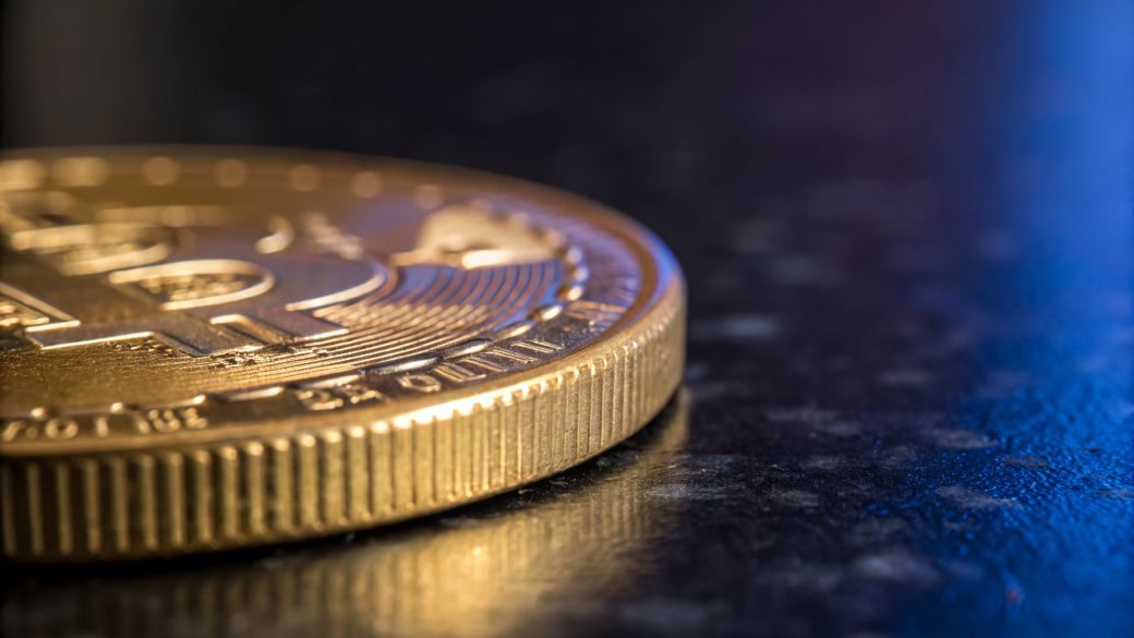 The image features a close-up of a gold-colored cryptocurrency coin, prominently displaying the Bitcoin symbol. The shiny surface of the coin reflects ambient light, creating a dynamic visual effect.