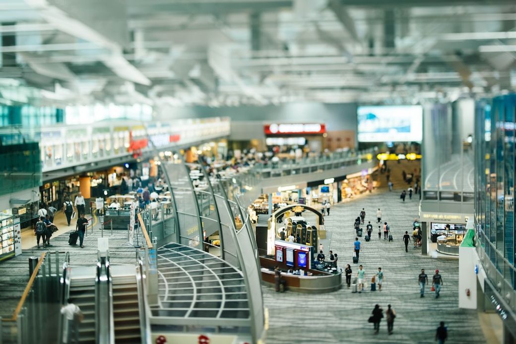This image captures a bustling airport terminal interior filled with travelers. The modern architecture features expansive shops and restaurants along the concourse. — Credit: Photo by CHUTTERSNAP on Unsplash