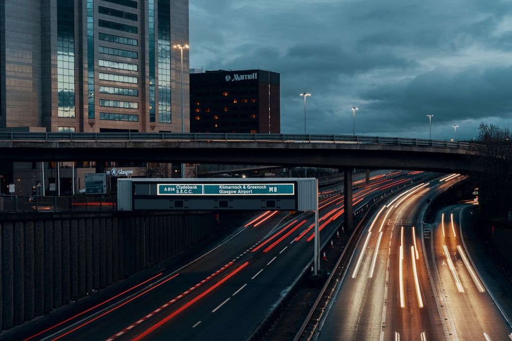 The image shows a highway overpass with light trails from moving vehicles at dusk. In the background, buildings such as a Marriott hotel and office structures are visible, along with signage directing to Glasgow Airport. — Credit: Photo by Ross Sneddon on Unsplash