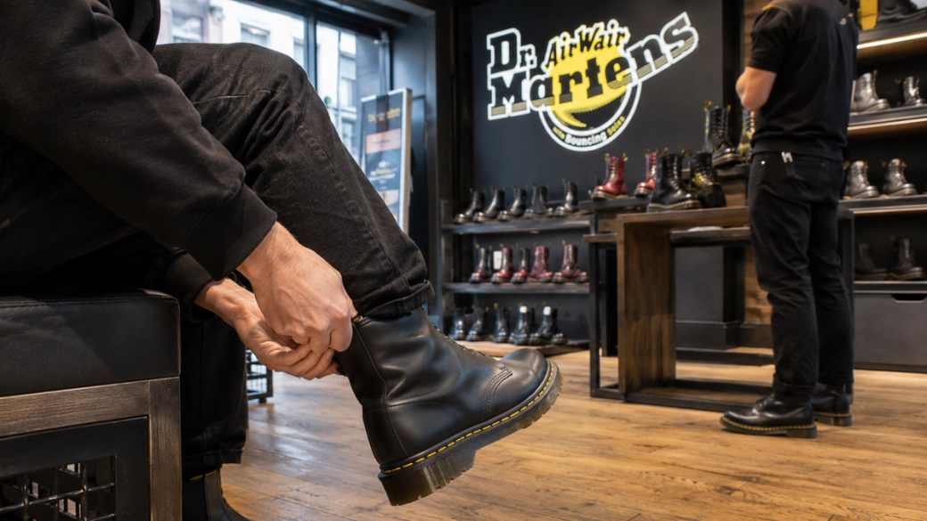 The image shows a person sitting on a bench in a shoe store, attempting to put on a black boot. In the background, various styles of Dr. Martens boots are displayed on shelves. Another individual is visible, standing while browsing the collection.