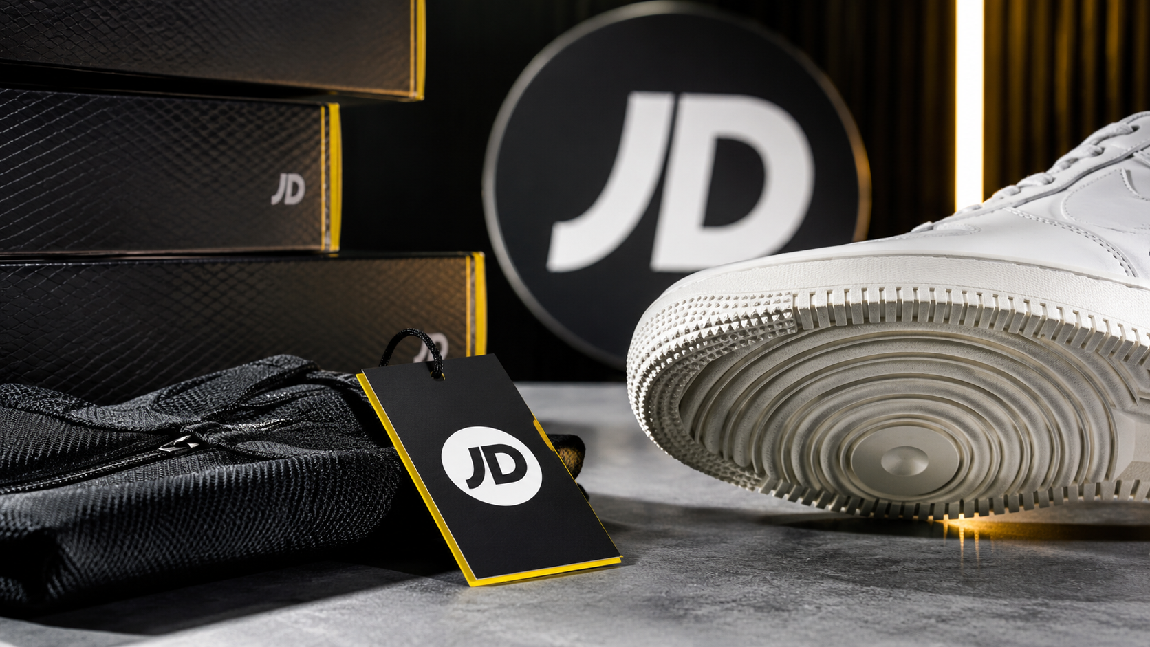 The image showcases a close-up of a white sneaker resting on a textured surface, with a prominent JD logo visible on a tag and in the background. Two dark boxes bearing the JD logo can also be seen, creating a focused brand presentation.