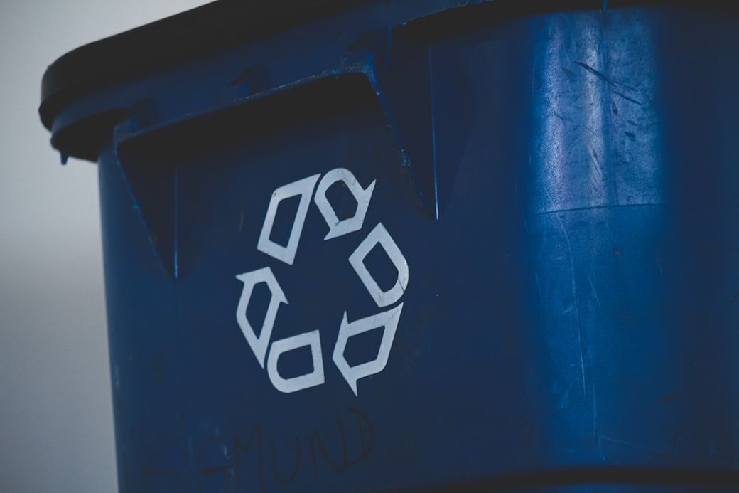 The image depicts a blue recycling bin with a visible recycling symbol on its side. The focus is on the bin's design and its purpose for waste separation. — Credit: Photo by Compagnons on Unsplash