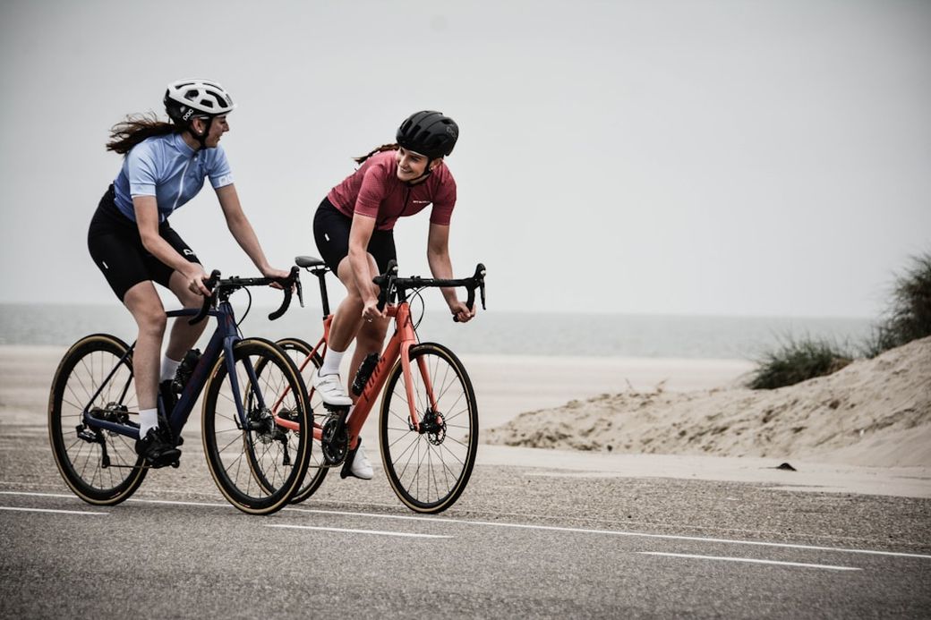 Two women are cycling along a coastal road with a beach in the background. They are engaged in a friendly conversation while riding their bikes. — Credit: Photo by Coen van de Broek on Unsplash