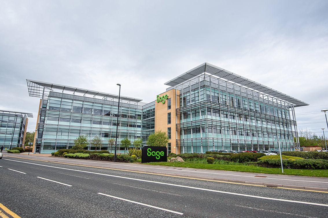 A modern glass office building prominently featuring the Sage brand. The structure is situated along a road, surrounded by well-maintained landscaping and green spaces.