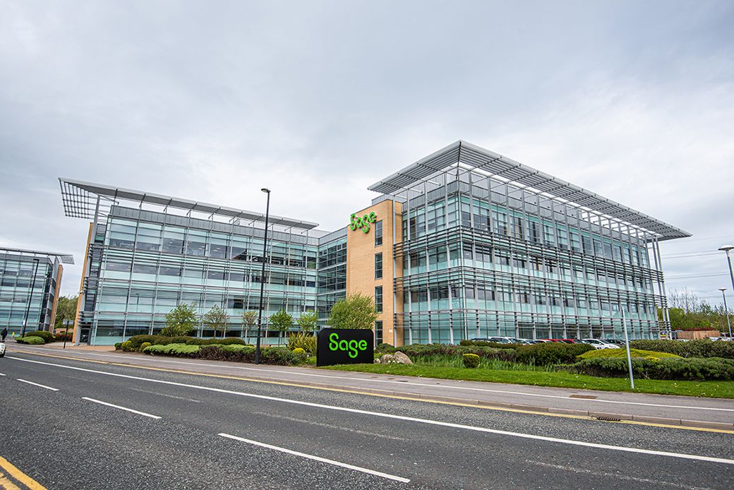 A modern glass office building prominently featuring the Sage brand. The structure is situated along a road, surrounded by well-maintained landscaping and green spaces.