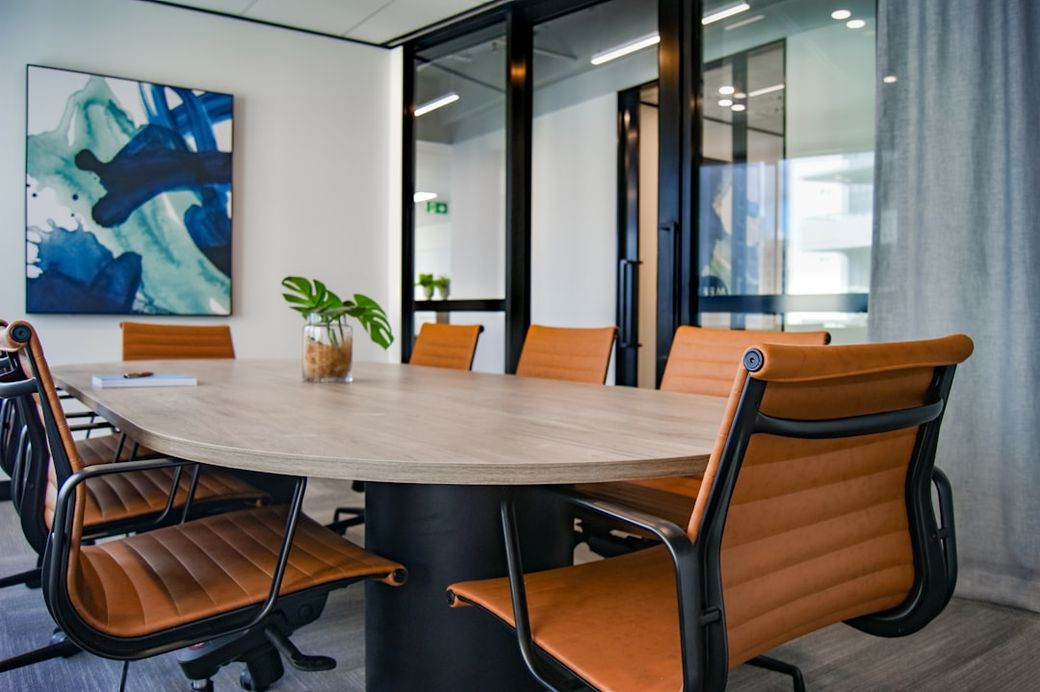 A modern conference room featuring an oval wooden table surrounded by orange leather chairs. The room has a contemporary design, with a piece of abstract art hanging on the wall and glass doors leading to another area. — Credit: Photo by S O C I A L . C U T on Unsplash