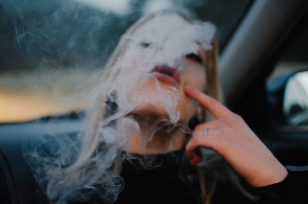 A close-up image of a woman seated in a car, with smoke swirling around her face. She appears to be engaging with the smoke, creating a mysterious and intimate atmosphere. — Credit: Photo by Duncan Shaffer on Unsplash