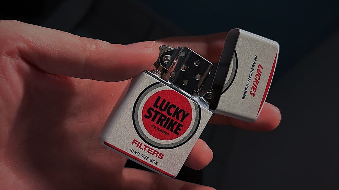 An individual holds a Lucky Strike cigarette pack with the flip-top lid open, displaying its bold red and white branding. The image highlights the product's design and aesthetic appeal.