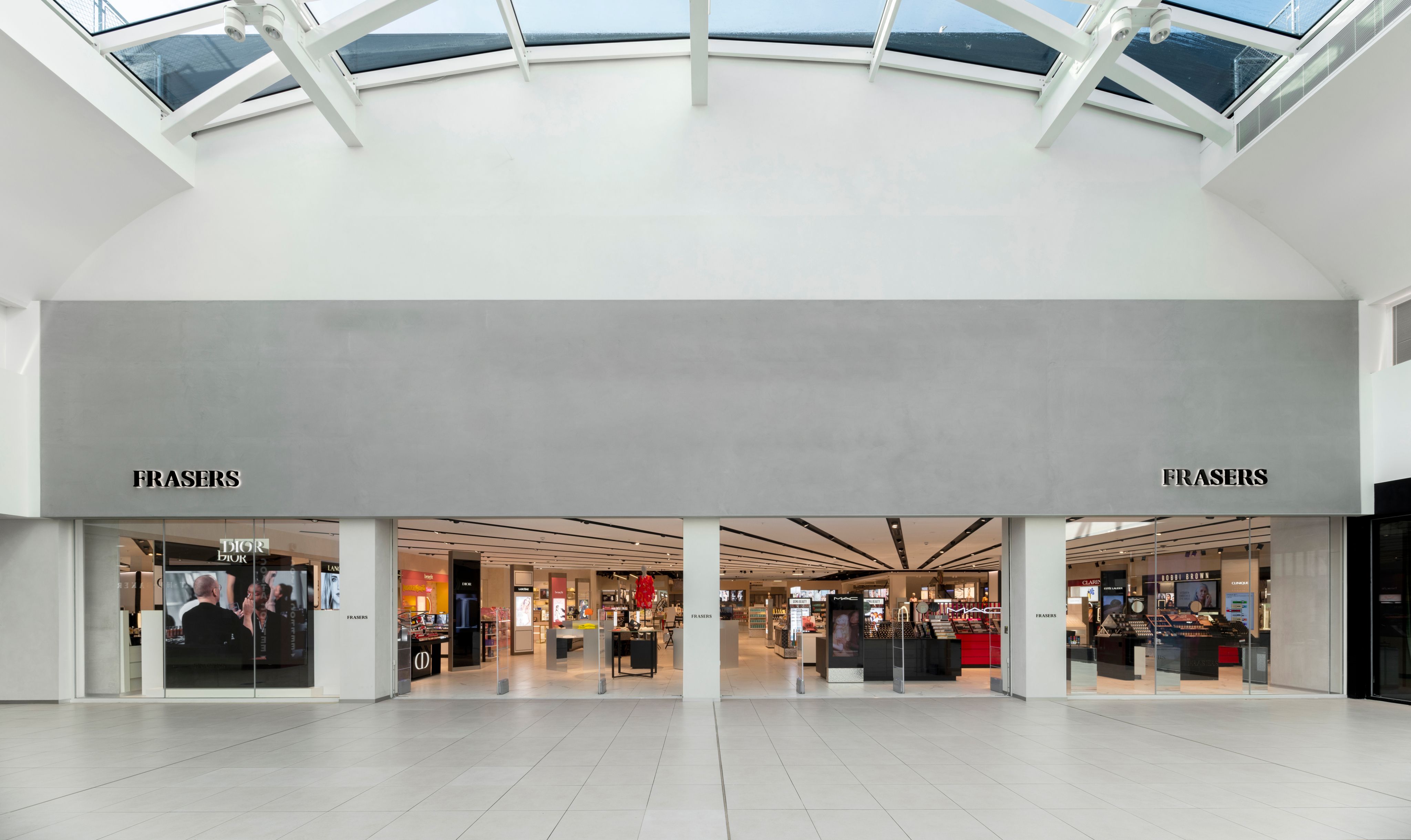 The image shows the entrance to a modern retail space with large glass doors leading to Frasers. The interior is well-lit with a minimalist design, featuring various retail displays visible through the glass.