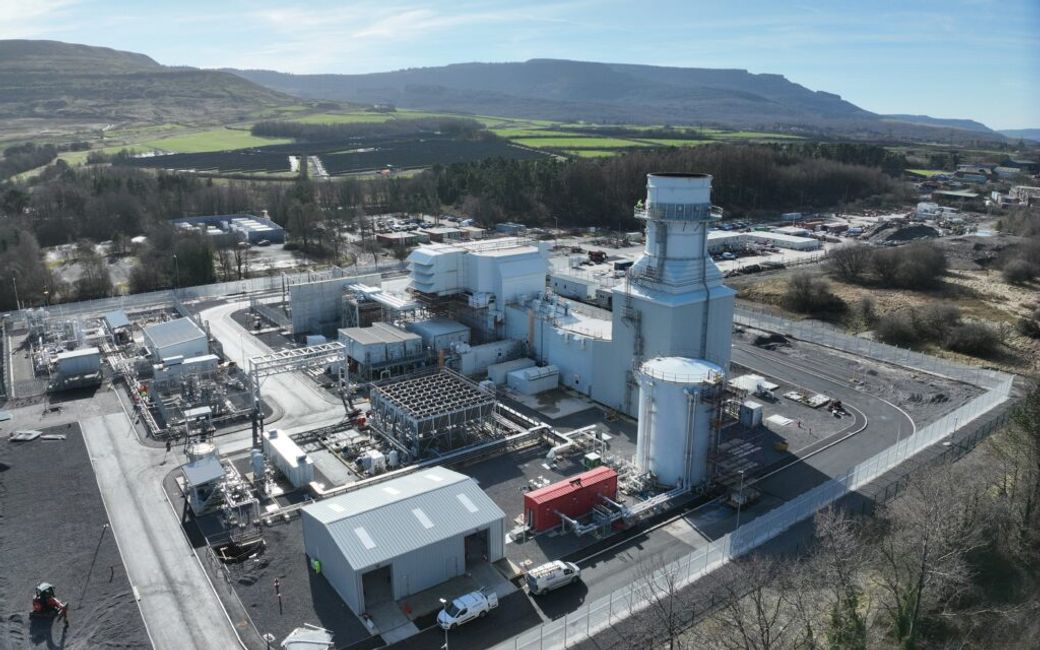 An aerial view of a modern gas power plant located in a rural setting. The facility features multiple structures with a large turbine building and associated infrastructure, surrounded by green hills and farmland.