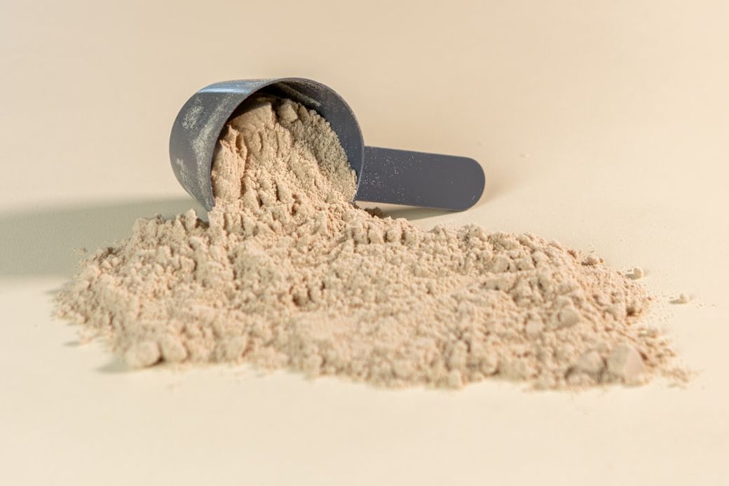 The image shows a measuring scoop tipped over, spilling a pile of light-colored powder onto a plain background. The fine texture and consistent color of the powder suggest it could be a protein supplement or baking ingredient. — Credit: Photo by Alex Saks on Unsplash