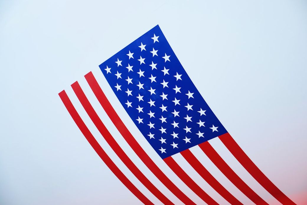 The image features a stylized representation of the American flag, featuring bold red stripes and a blue rectangular field adorned with white stars. The flag appears to be tilted, creating a dynamic visual effect. — Credit: Photo by Brian McGowan on Unsplash
