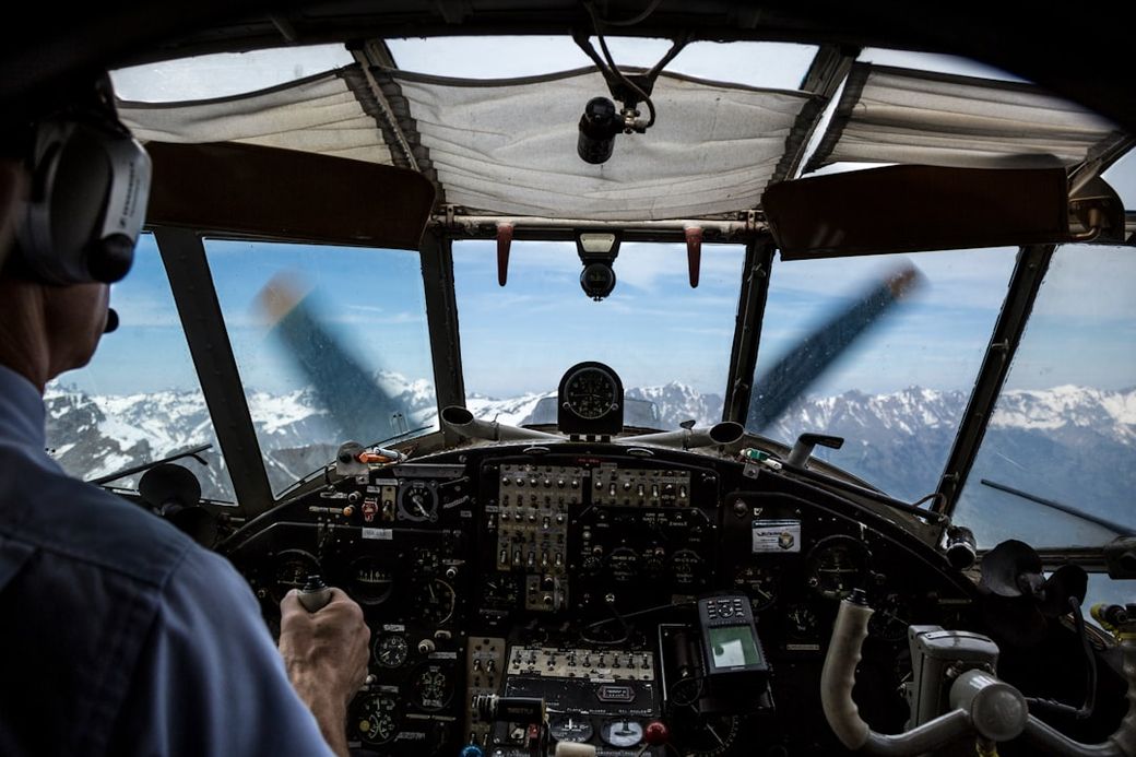 The cockpit of a vintage aircraft is shown, with a pilot actively controlling the plane while flying over snow-capped mountains. The dashboard is filled with various gauges and controls, showcasing a view of the sky and mountains through the cockpit windows. — Credit: Photo by Laurent Perren on Unsplash