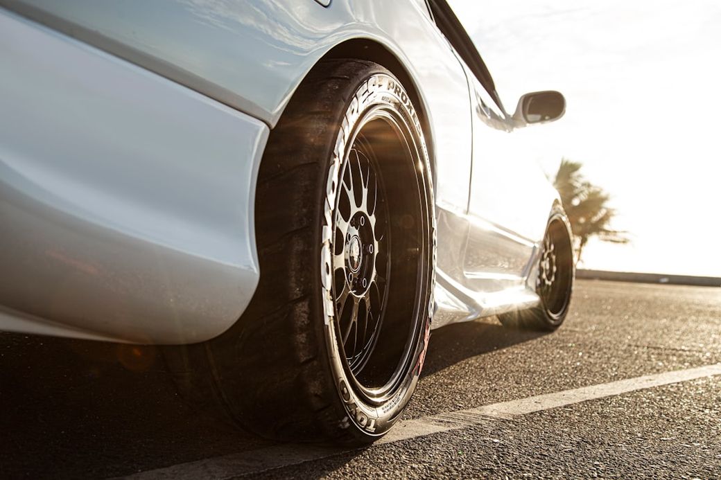 The image depicts a close-up view of a modified car, focusing on its sleek, low profile and distinctive wheel design. The sun is setting in the background, casting warm light across the scene. — Credit: Photo by Velito on Unsplash