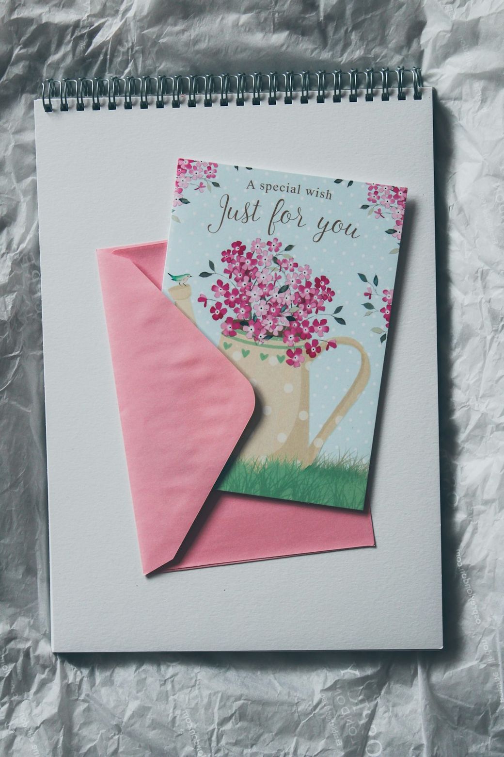 The image features a decorative greeting card with a floral design resting on top of a blank spiral notebook. The card is slightly opened, revealing a pink envelope underneath. — Credit: Photo by Siora Photography on Unsplash