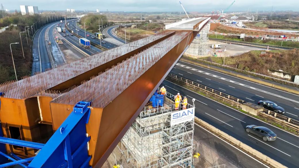 The image captures a construction site where a large steel beam is being positioned over a highway. Workers can be seen near the beam, which is part of an infrastructure project, with scaffolding and cranes in the background.