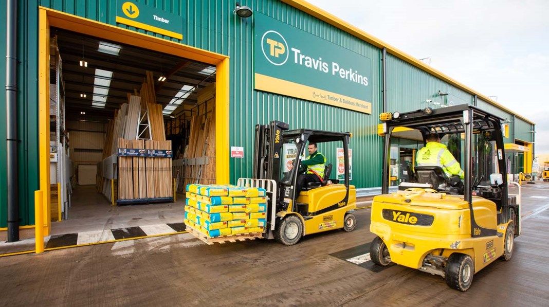 The image depicts a bustling scene outside a Travis Perkins building materials store. Two forklift trucks are in operation, one unloading stacked materials while the other is parked at the entrance.