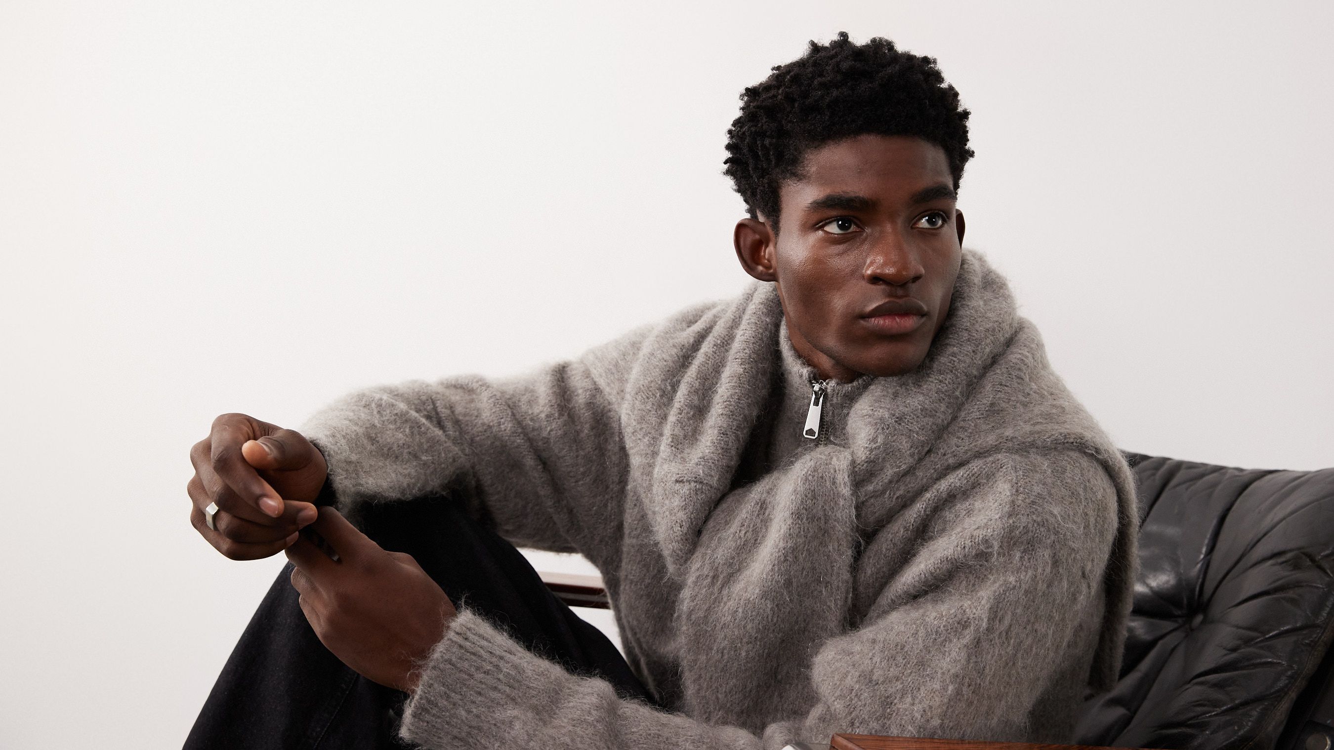 A model sits casually on a chair, wearing a cozy gray knit sweater. The minimalist background emphasizes the stylish garment and the model's relaxed pose.