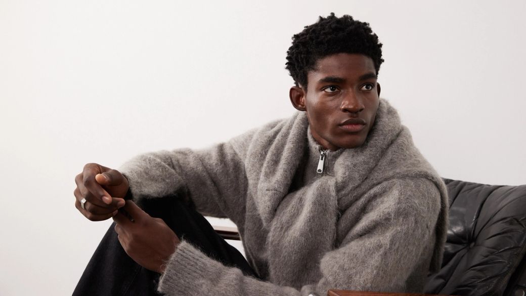 A model sits casually on a chair, wearing a cozy gray knit sweater. The minimalist background emphasizes the stylish garment and the model's relaxed pose.