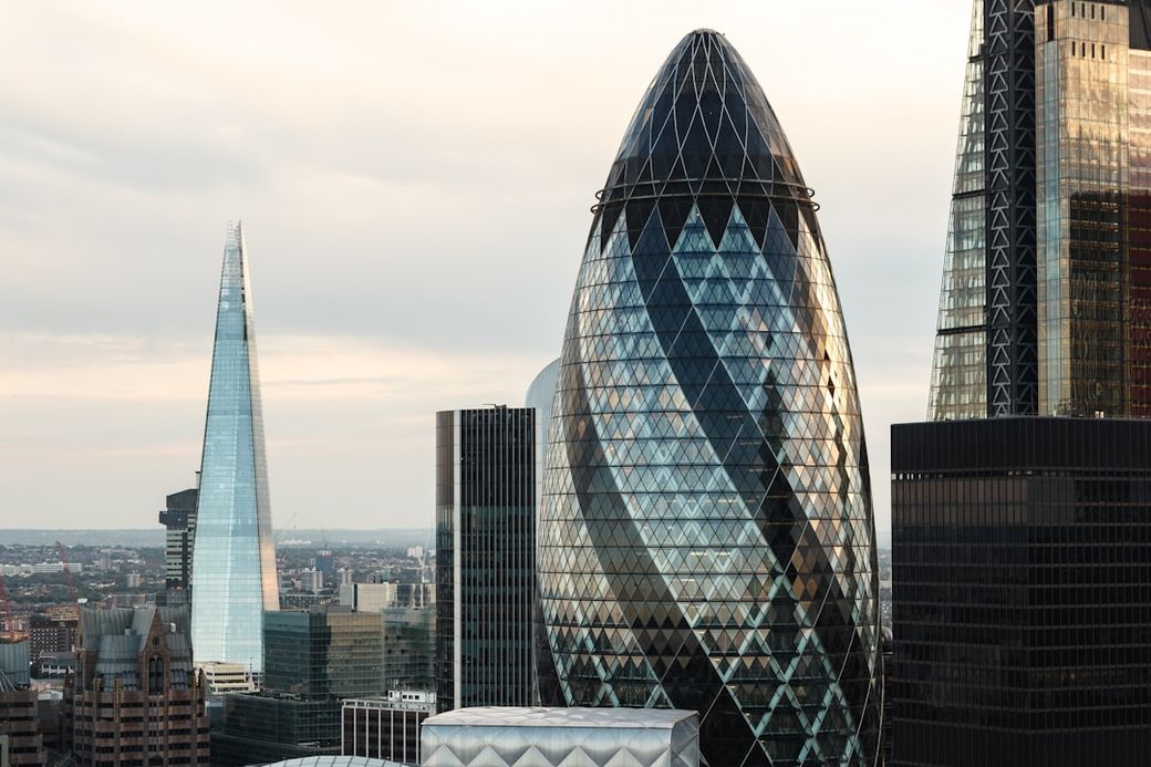 The image captures a skyline view of modern skyscrapers in London, featuring notable buildings like the Gherkin and The Shard. The reflections on the glass surfaces contrast against the cloudy sky, showcasing contemporary architectural design. — Credit: Photo by Ed Robertson on Unsplash