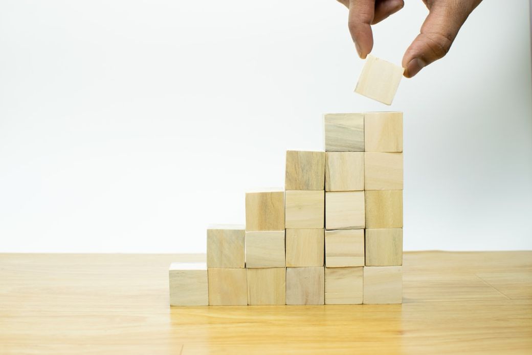 A hand placing a wooden block atop a stepped arrangement of similar blocks. The stacked blocks create a visual representation of growth or progress, set against a plain white background. — Credit: Photo by Imagine Buddy on Unsplash