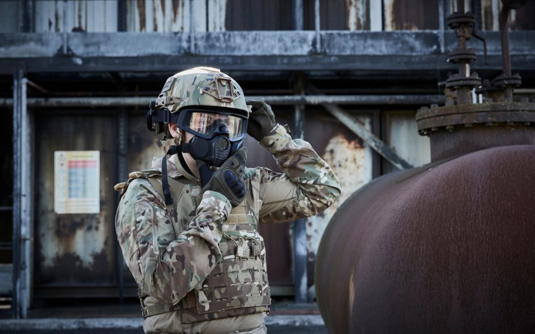 A soldier in camouflage gear adjusts his protective mask while standing in an industrial setting. The background features rusted structures, indicating a potentially hazardous environment.
