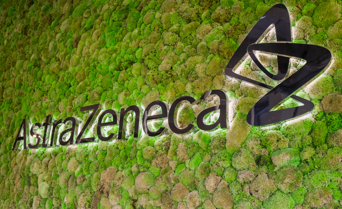The image showcases a green moss-covered wall featuring the logo of the pharmaceutical company AstraZeneca. The natural elements create a calming environment that reflects a modern and eco-friendly design approach.