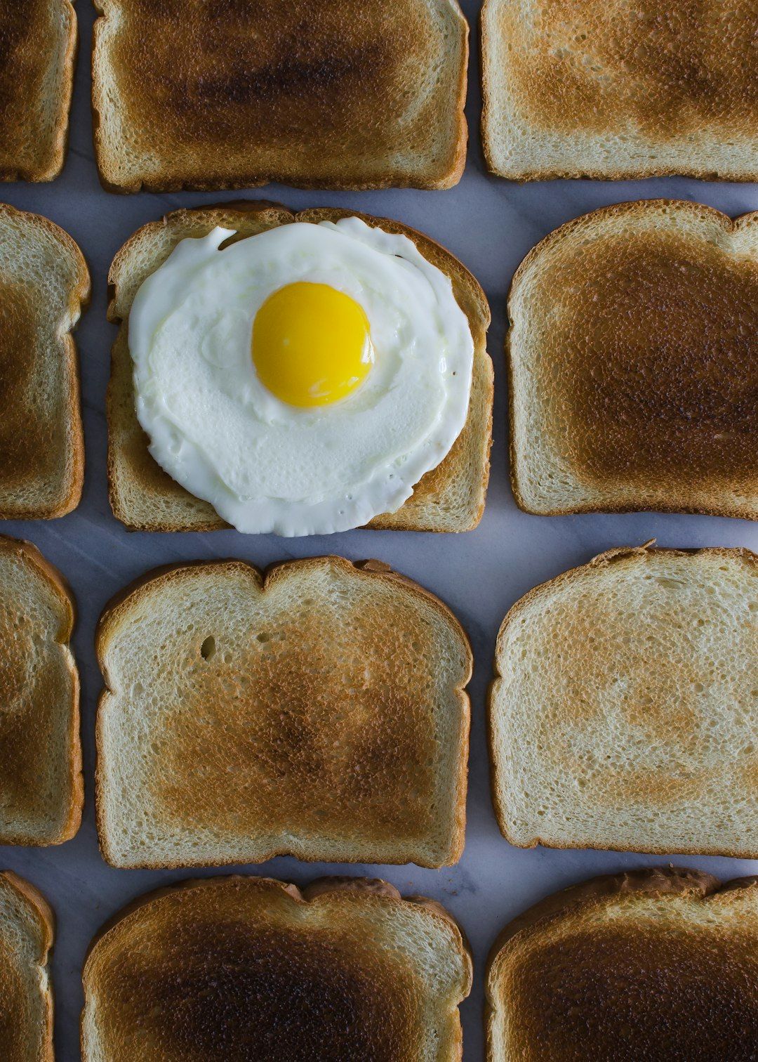 The image features several slices of toasted bread arranged in a grid pattern, with one slice topped by a fried egg. The highlighted egg contrasts with the surrounding toast, creating a visually striking focal point.