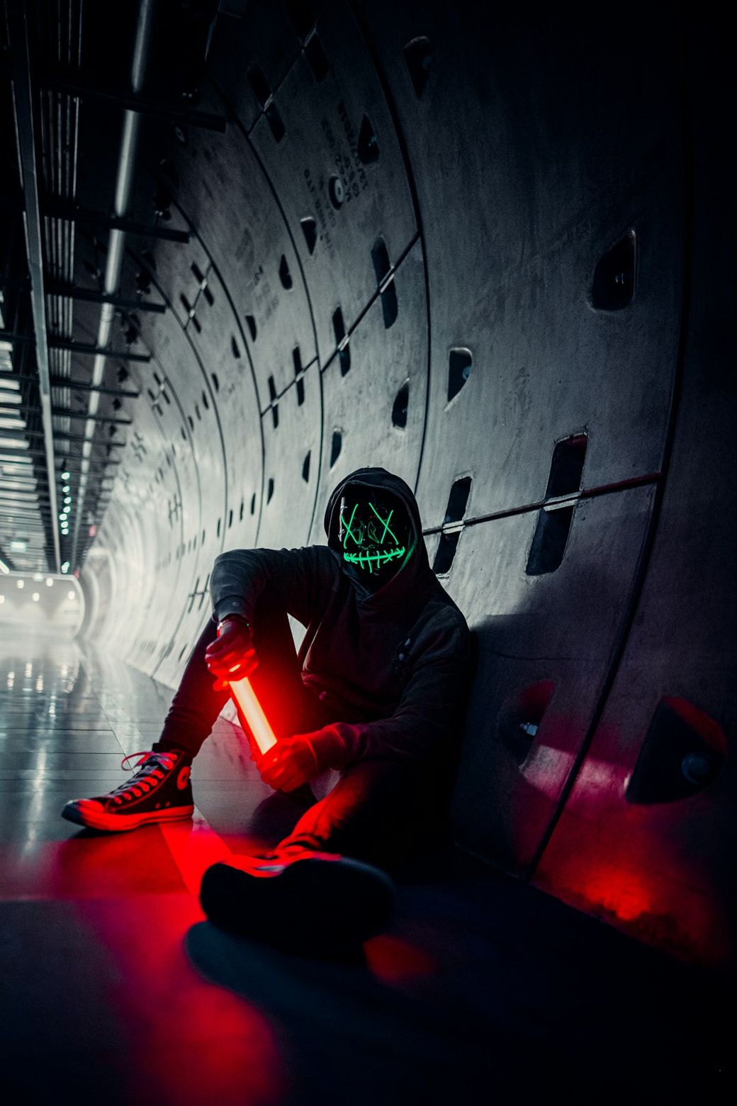 A person sits against a curved wall in a dimly lit tunnel, wearing a hood and a mask that glows green with a skull design. They hold a red light, creating a striking contrast with the dark surroundings. — Credit: Photo by Joshua Rawson-Harris on Unsplash