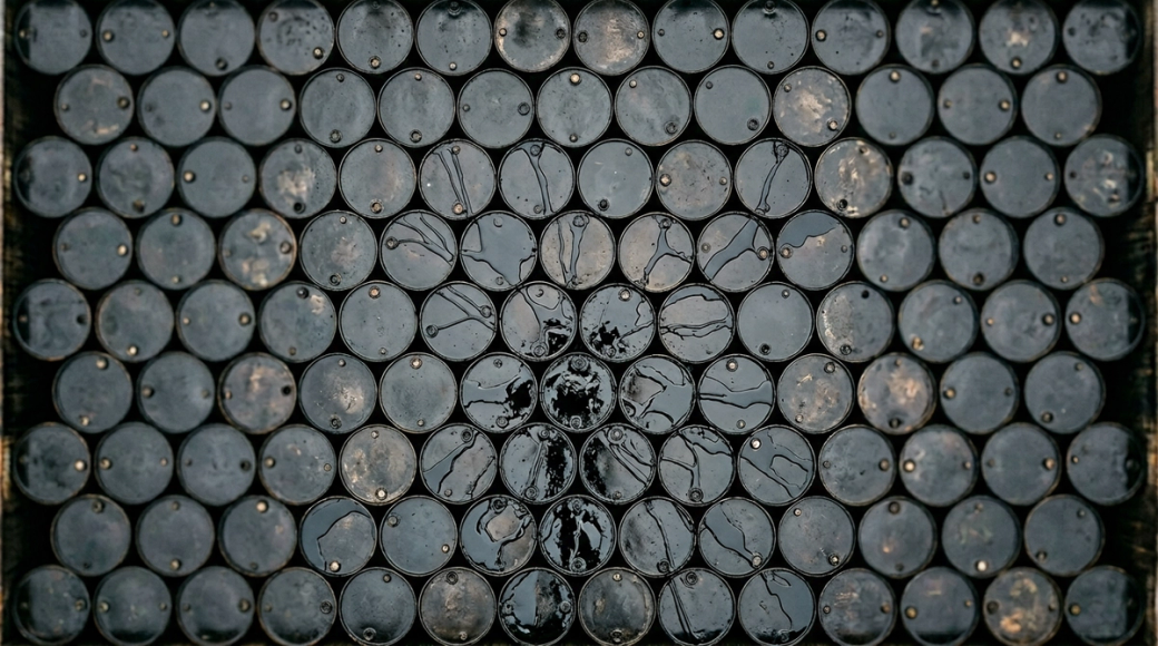 The image showcases a tightly packed arrangement of oil drums, all filled to capacity, resembling a black industrial surface. Crude oil overflows from the rims of the drums, creating a network of glossy tributaries that connect them, evoking a sense of surplus and market saturation.