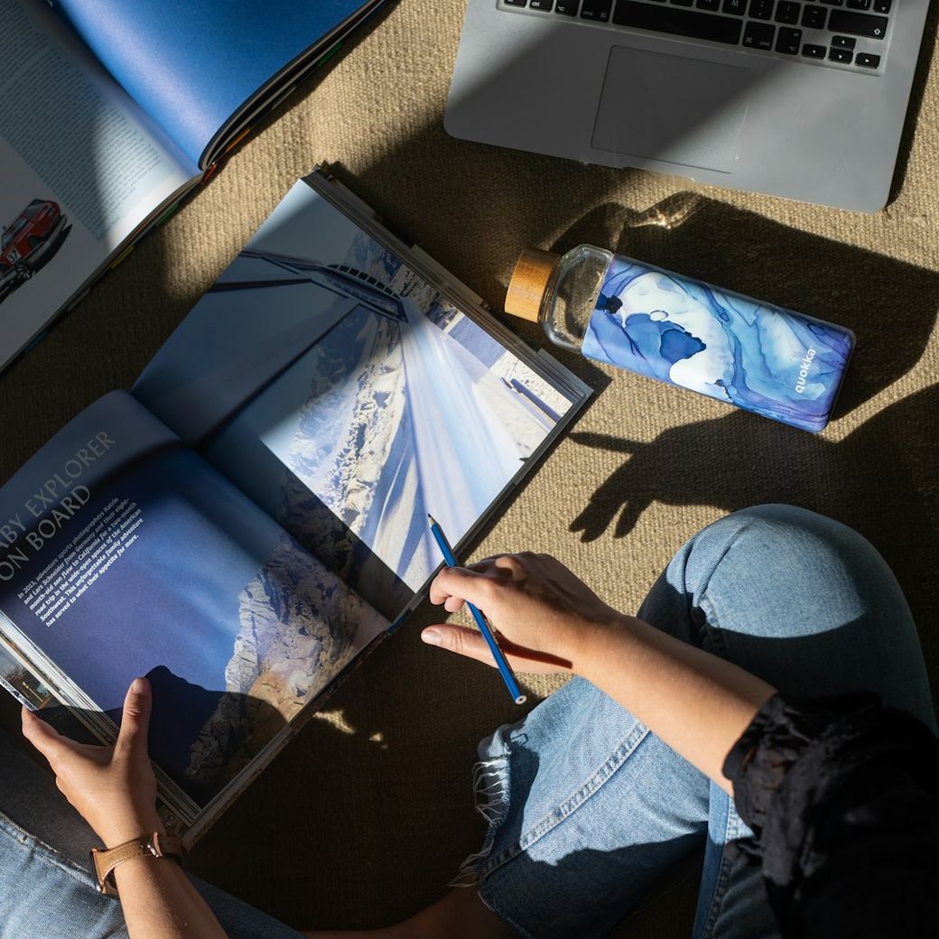 A person seated on a floor, reviewing a travel magazine or guide featuring scenic landscapes. Nearby, a laptop is visible, along with a water bottle, suggesting a casual yet focused atmosphere. — Credit: Photo by quokkabottles on Unsplash