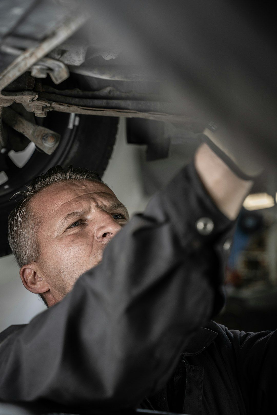 A mechanic is working underneath a vehicle, focusing intently on a task. The image captures the detailed environment of an auto repair shop with tools and equipment in the background.
