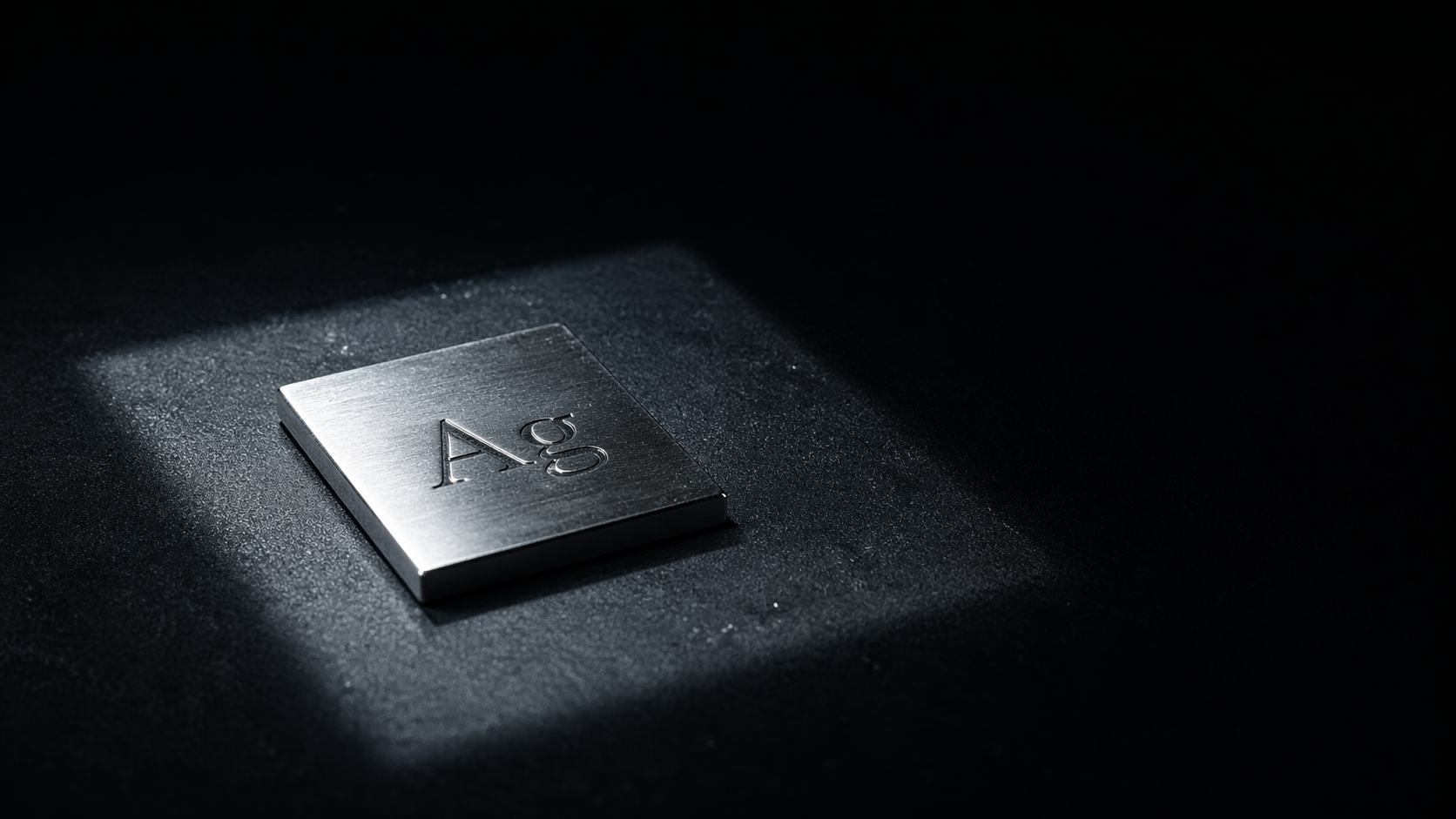 The image features a shiny metal plate embossed with the chemical symbol 'Ag', representing silver. It is set against a dark background, illuminated by a narrow beam of light, highlighting the reflective surface of the metal.