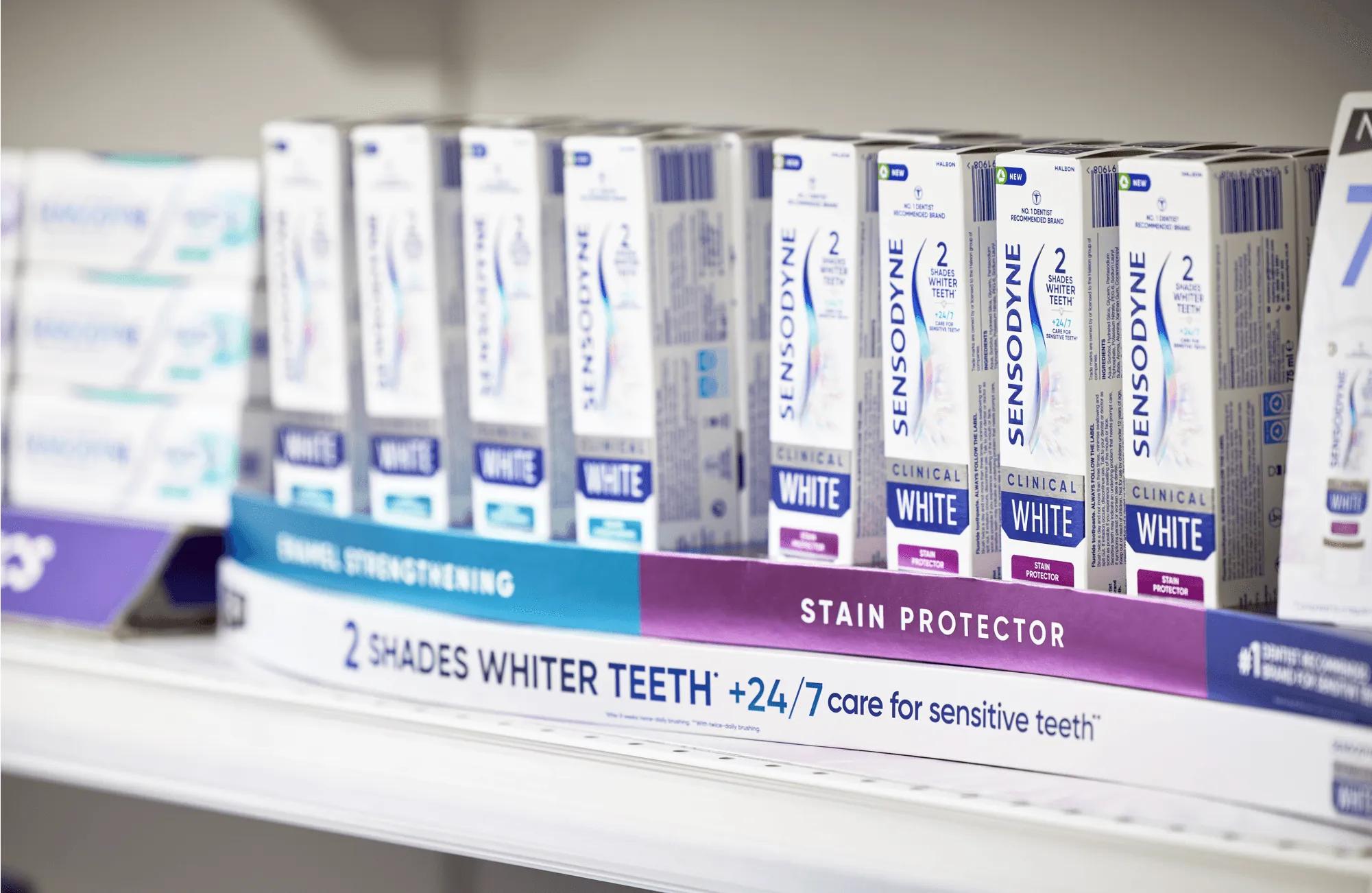 The image shows a shelf filled with various packages of Sensodyne toothpaste, prominently displaying multiple boxes with distinct branding and product details. The setting appears to be a retail space, specifically in the personal care or oral hygiene section.