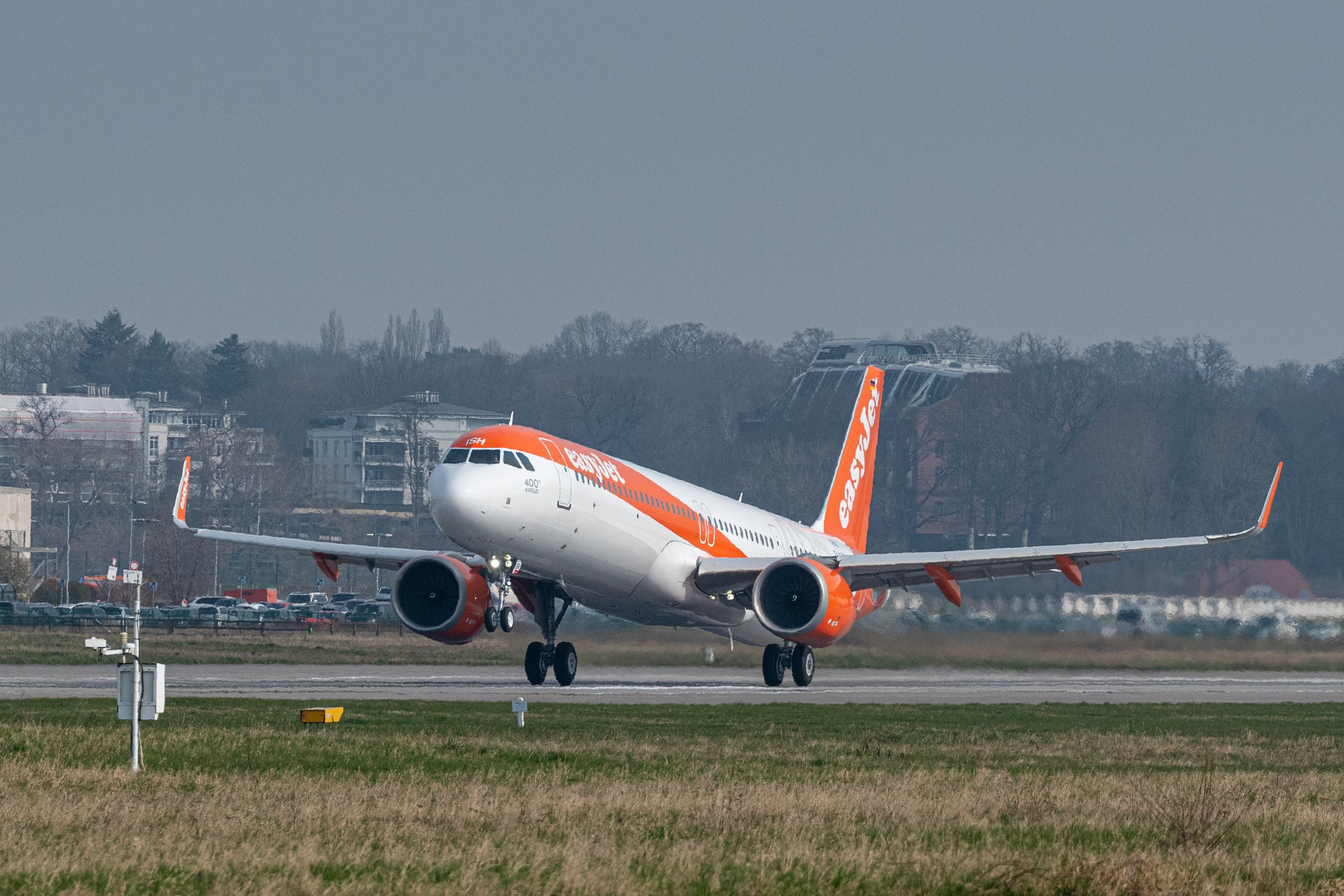 An EasyJet aircraft is taking off from a runway, lifting off the ground with its wheels in the air. The background features a mix of urban buildings and trees under a clear sky.