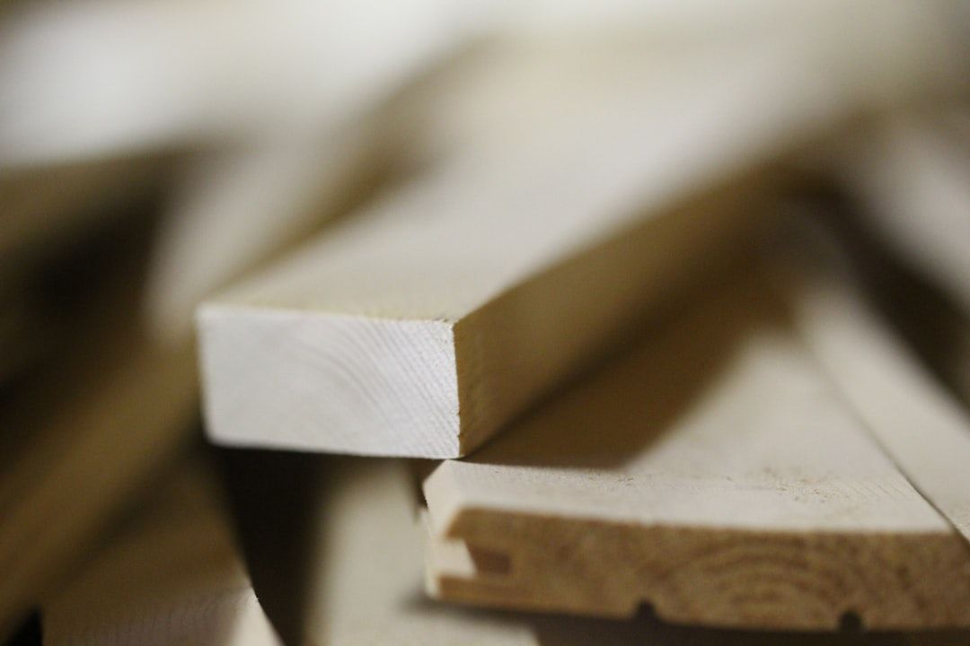 The image shows a close-up view of a stack of wooden planks, highlighting the texture and grain of the wood. The focus is on an individual plank in the foreground, with others slightly blurred in the background.