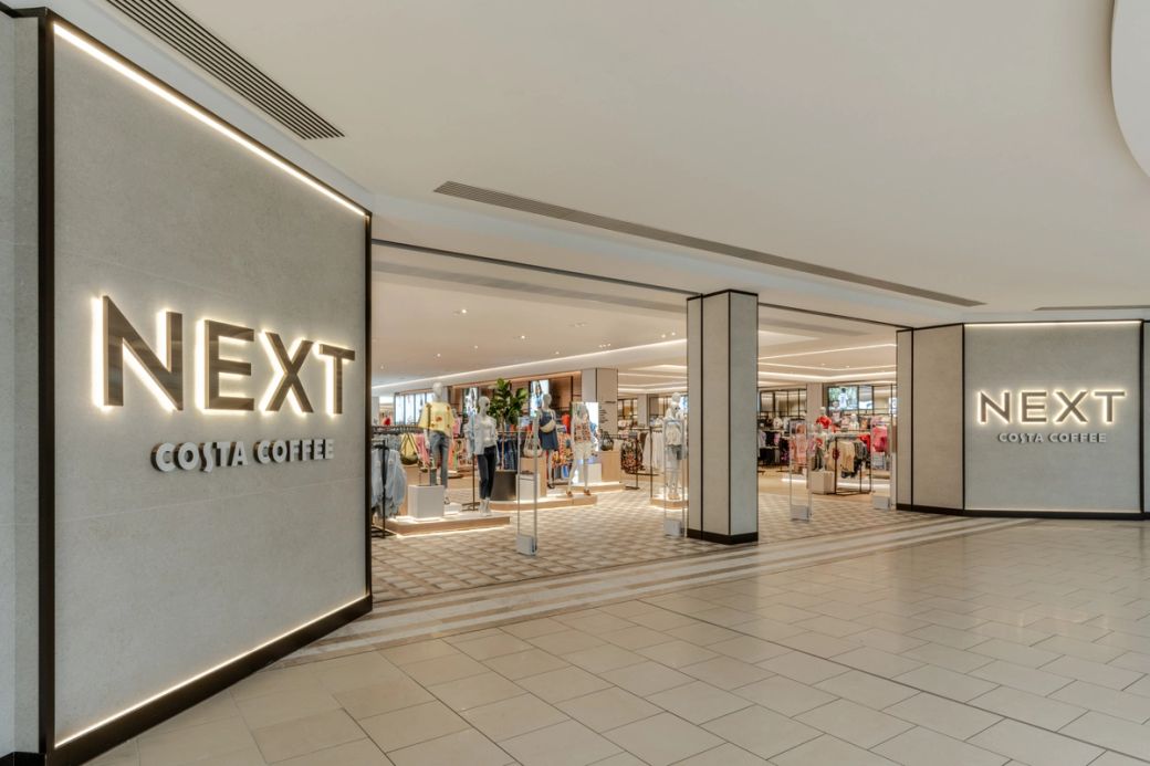 The image shows the interior of a retail shopping area featuring the storefronts of 'NEXT' and 'COSTA COFFEE'. The design is modern with bright lighting and displays of apparel visible inside.