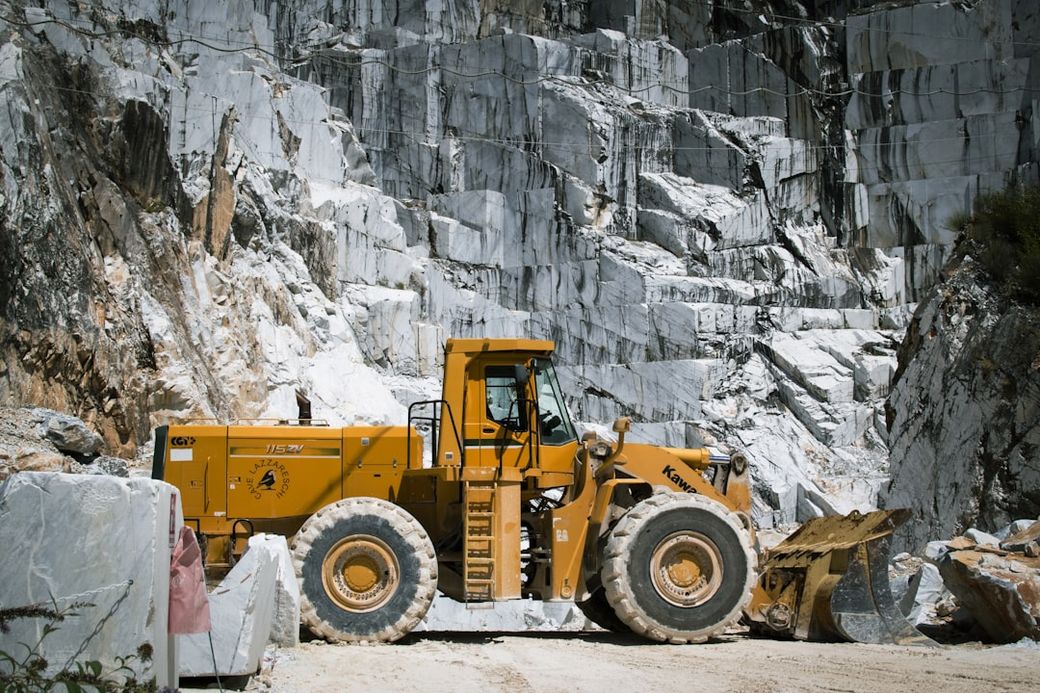 The image shows a yellow heavy machinery loader positioned near a marble quarry. The backdrop features large, rocky marble surfaces, indicating an active quarrying operation. — Credit: Photo by Gianluigi Marin on Unsplash