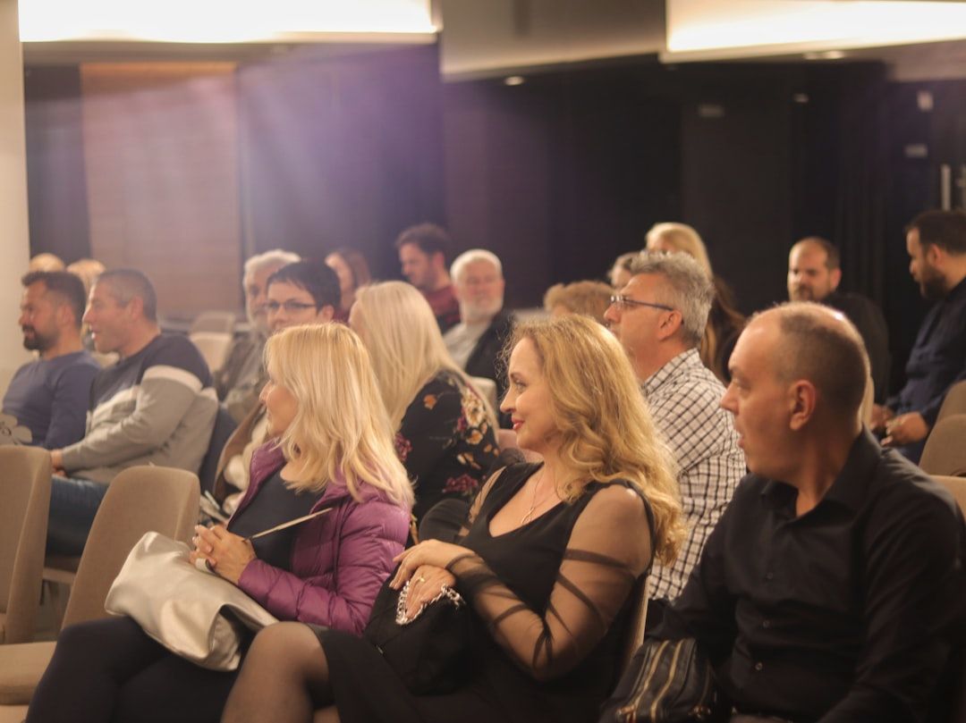 A diverse audience is seated in a modern conference setting, attentively listening to a speaker or presentation. The room is well-lit, showcasing a mix of individuals engaged in the event.