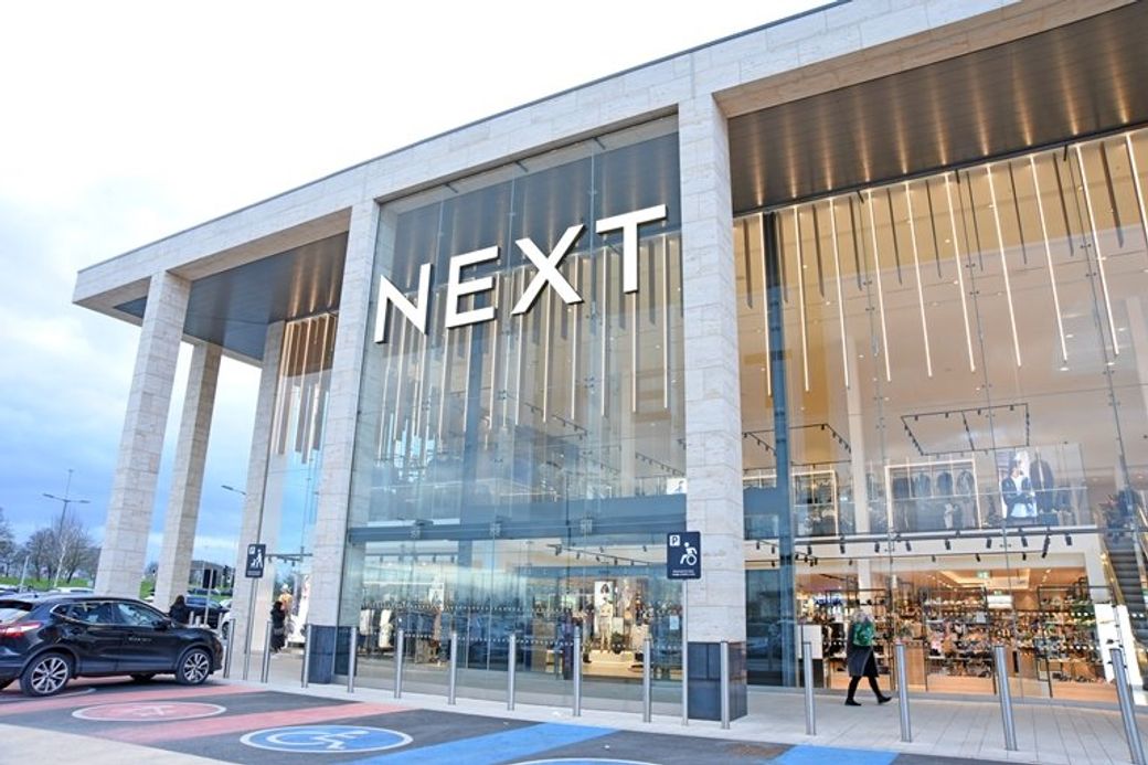 The image shows the exterior of a NEXT retail store with modern architectural features. The store is prominently displayed with large glass windows and a clean design, reflecting a contemporary shopping environment.
