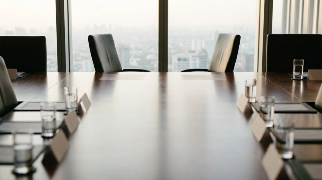 The image depicts a long boardroom table set for an upcoming meeting, with leather chairs neatly arranged around it. Two chairs at one end are pulled back and appear vacant, suggesting a recent reorganization or transition, while the other chairs remain in place with name placards and glasses of water, illuminated by warm afternoon light filtering through tall windows.