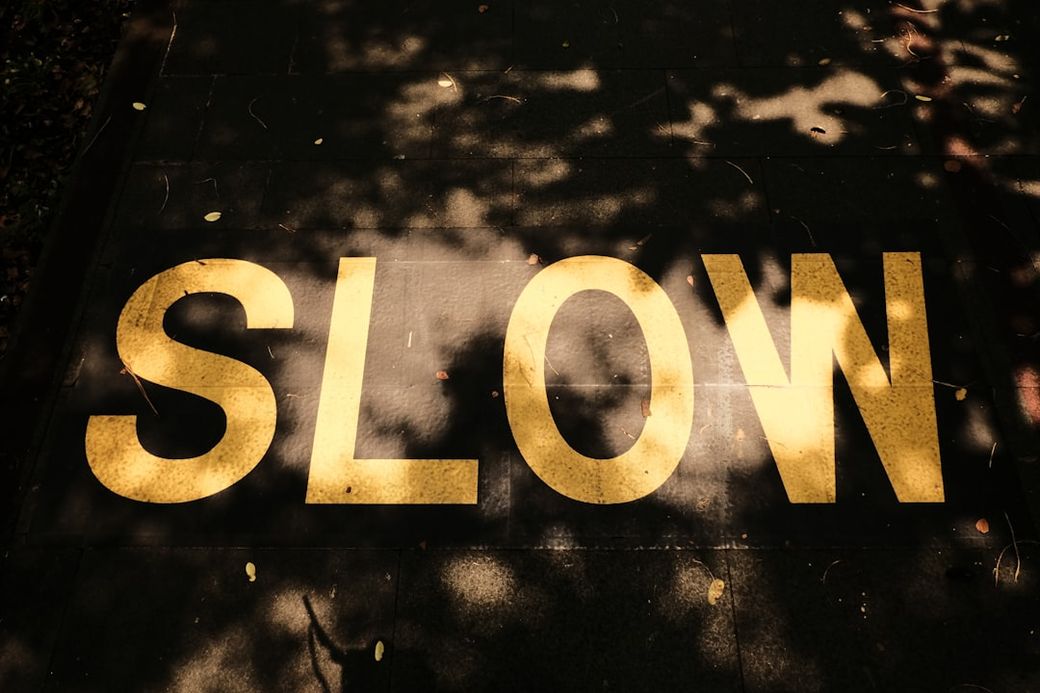 The image shows a large, yellow, capitalized word 'SLOW' painted on a dark pavement. The area is partly shaded, creating a contrast between the bright lettering and the darker background. — Credit: Photo by Anisa Gauri on Unsplash