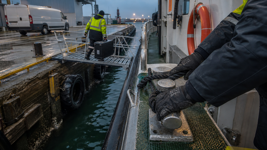 The image shows workers on a docked vessel preparing for operation. One worker is adjusting equipment while another is in the background by a loading ramp, suggesting maritime logistics activity.