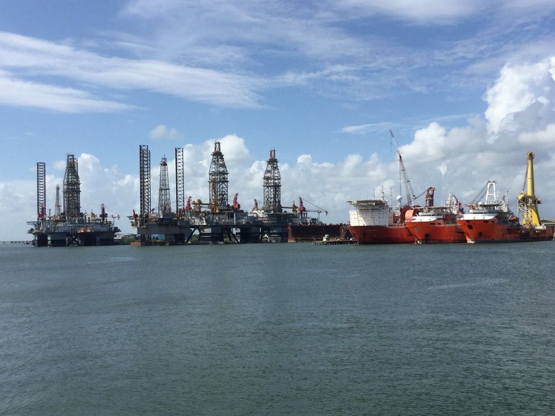 The image depicts a busy offshore oil rig facility with multiple drilling platforms and support ships operating in calm waters under a blue sky with scattered clouds. The installation showcases the industrial scale and complexity of offshore oil extraction.