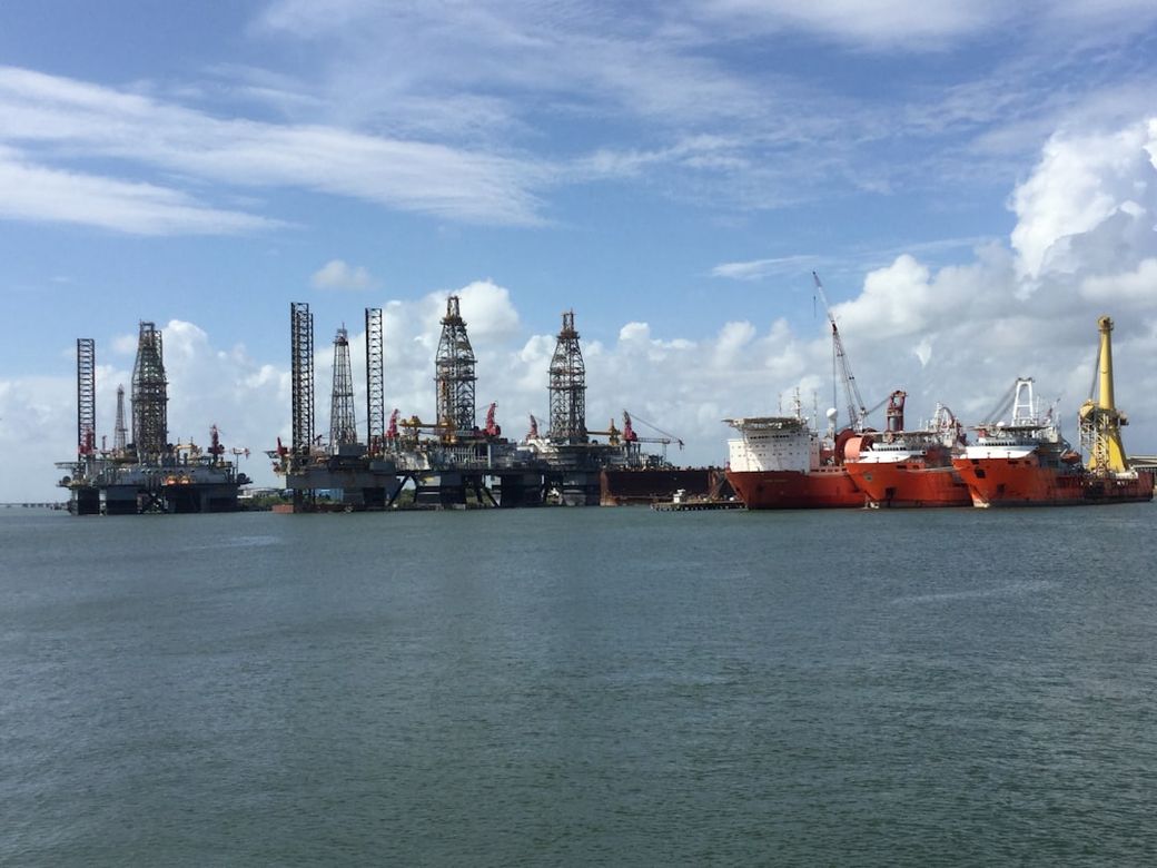 The image depicts a busy offshore oil rig facility with multiple drilling platforms and support ships operating in calm waters under a blue sky with scattered clouds. The installation showcases the industrial scale and complexity of offshore oil extraction. — Credit: Photo by Linda Finkin on Unsplash