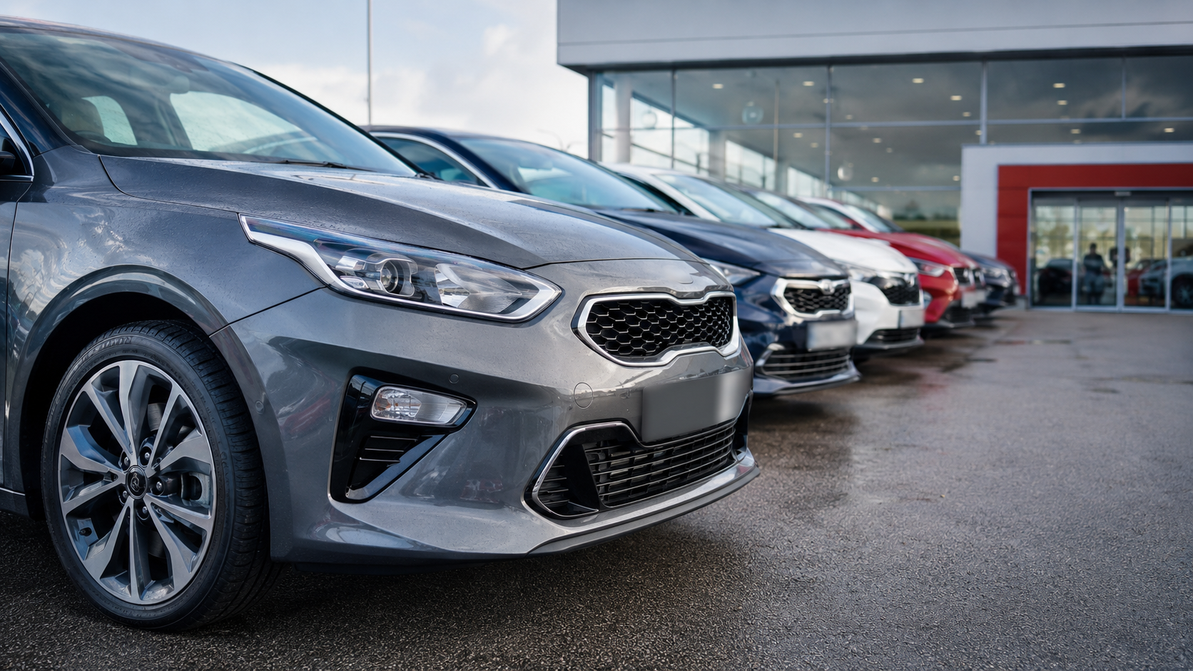 A row of various cars lined up outside a modern car dealership. The foreground features a prominently displayed gray vehicle, while multiple other cars in different colors are visible in the background.