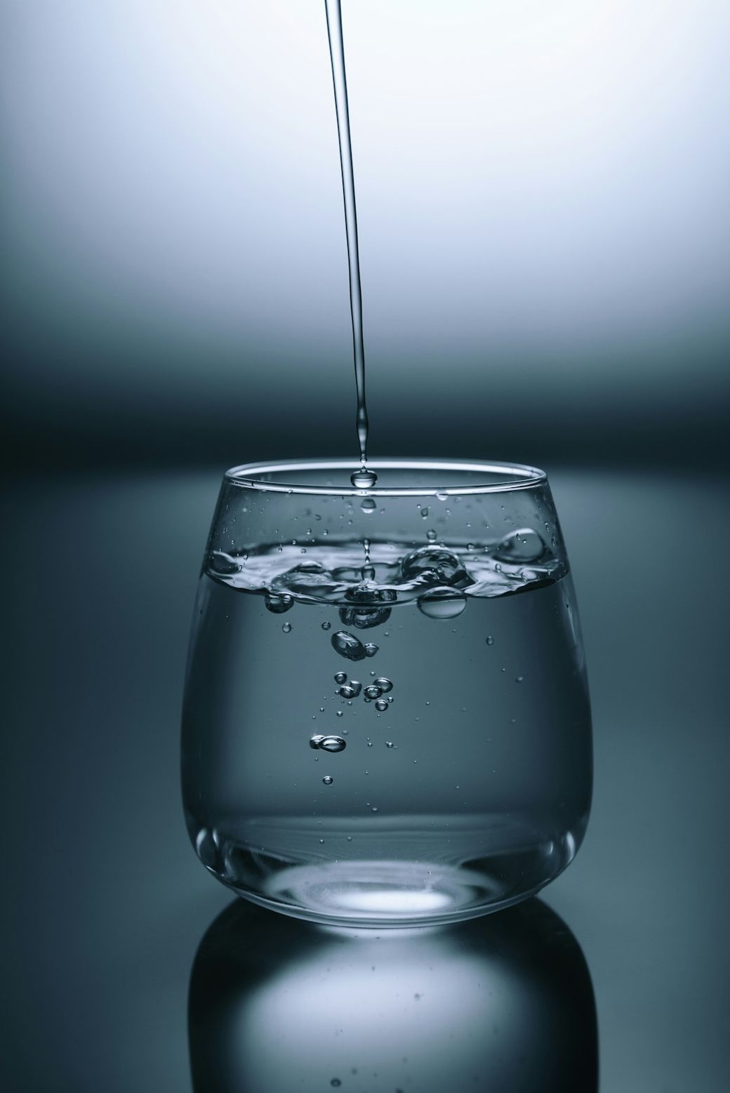 The image depicts a clear glass filled with water, with droplets and air bubbles visible. Water is being poured into the glass, creating ripples on the surface. — Credit: Photo by KOBU Agency on Unsplash