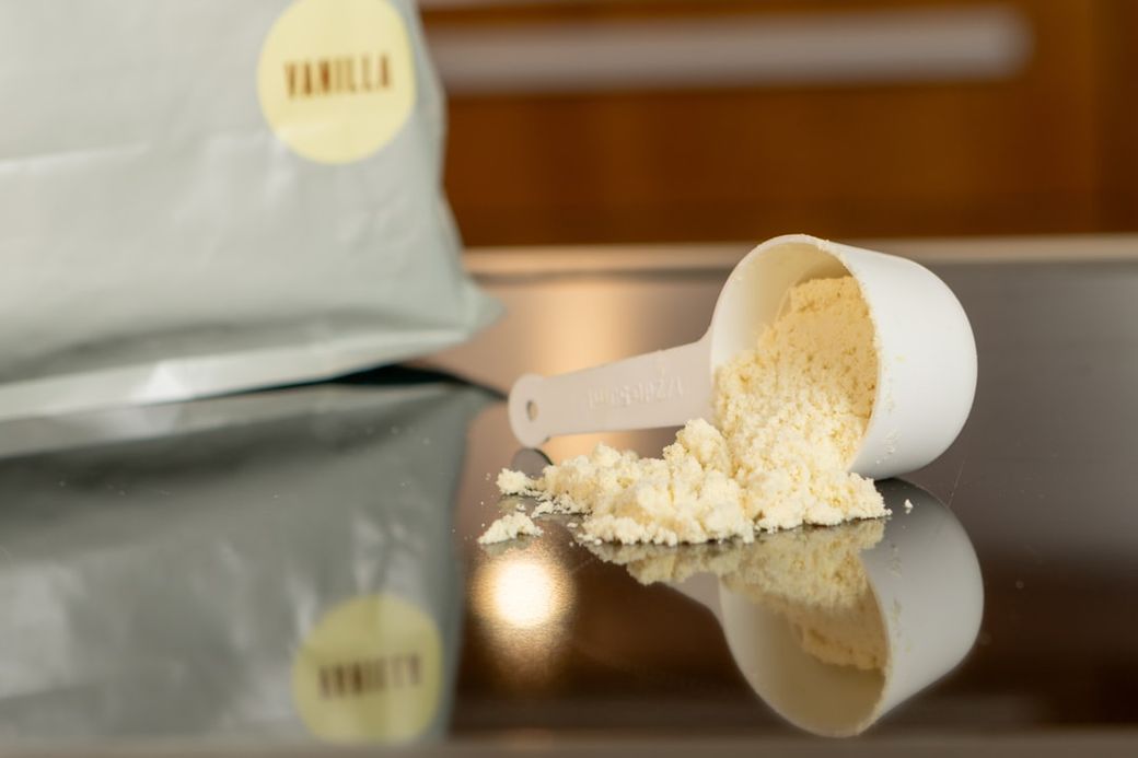 The image shows a white scoop measuring device next to a spilled portion of vanilla powder on a reflective surface. A partially visible bag of vanilla powder can be seen in the background. — Credit: Photo by Alex Saks on Unsplash