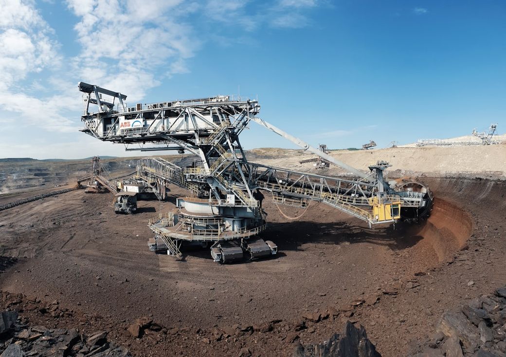 The image depicts a large bucket-wheel excavator operating in an open-pit mining site, set against a clear blue sky. The machinery is surrounded by earthy terrain, showcasing the scale of industrial mining operations. — Credit: Photo by Albert Hyseni on Unsplash
