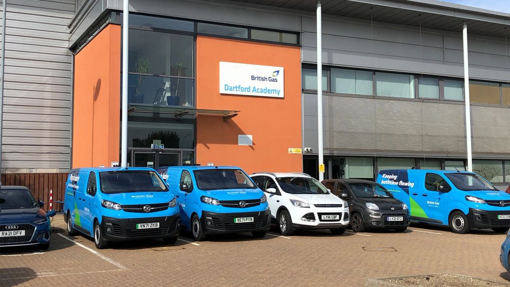 The image shows the exterior of Dartford Academy, featuring blue delivery vans parked outside. The building has large glass windows and a prominent sign that identifies it as the Dartford Academy.
